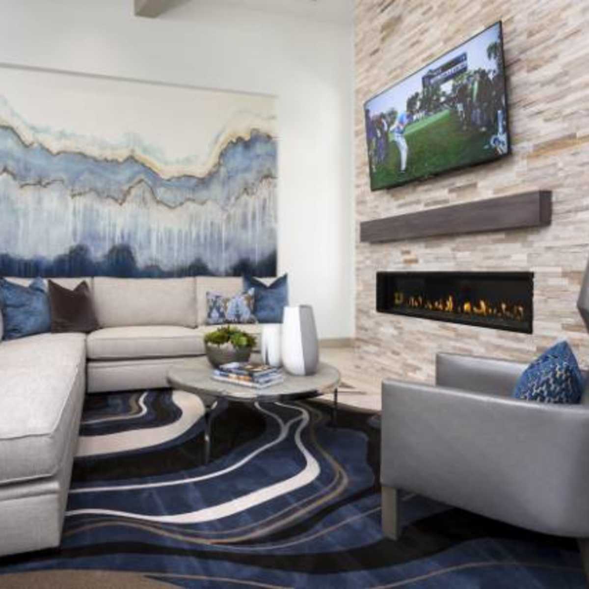 A modern living room with a sectional sofa, a wall-mounted television, and a stone fireplace, all complemented by abstract artwork and a patterned area rug.