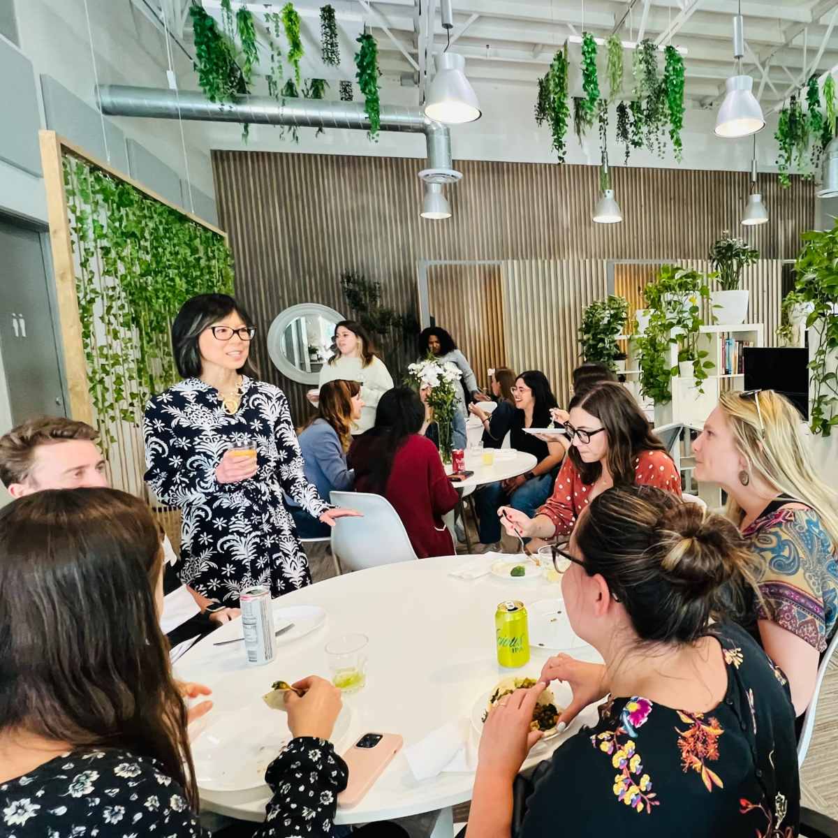 A group of people gathers around a table in a bright, plant-decorated office space, sharing food and conversation.