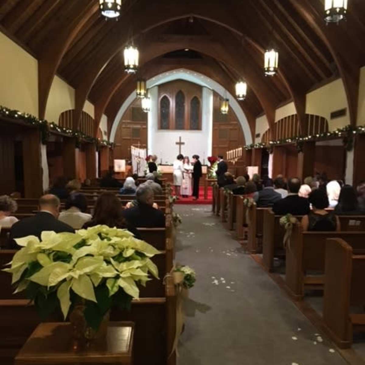 A wedding ceremony takes place inside a church, with guests seated in wooden pews and decorations featuring greenery and white flowers.