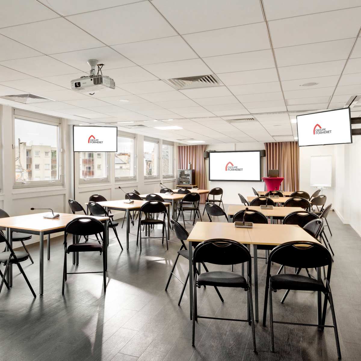 A modern classroom setup features several tables and chairs arranged for a presentation, with large screens displaying a logo on the walls.