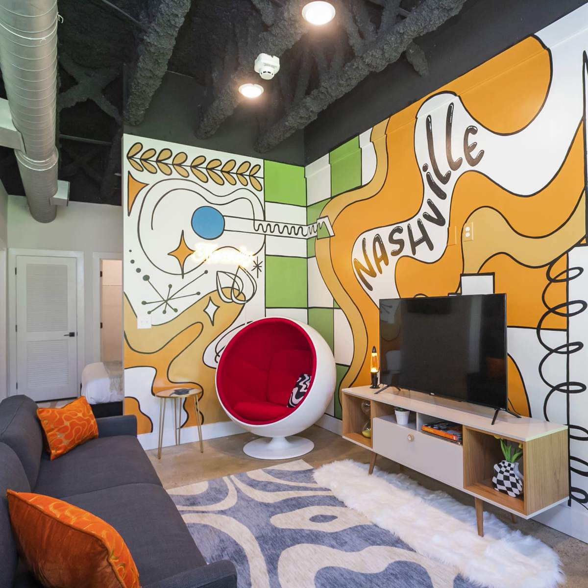 The room features a colorful mural on the walls, a modern white chair, a television stand, and a sofa with orange accents.