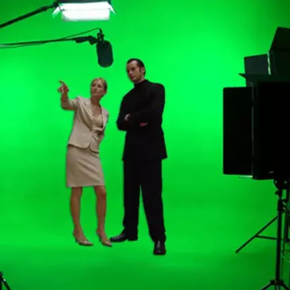A woman in business attire and a man in black clothing stand on a green screen set, surrounded by studio lighting equipment.