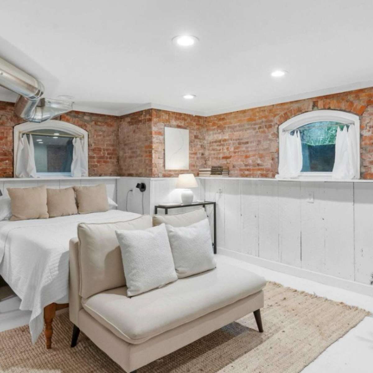 A bright basement room featuring a bed with neutral bedding, a small sofa, and exposed brick walls with white-paneled accents.