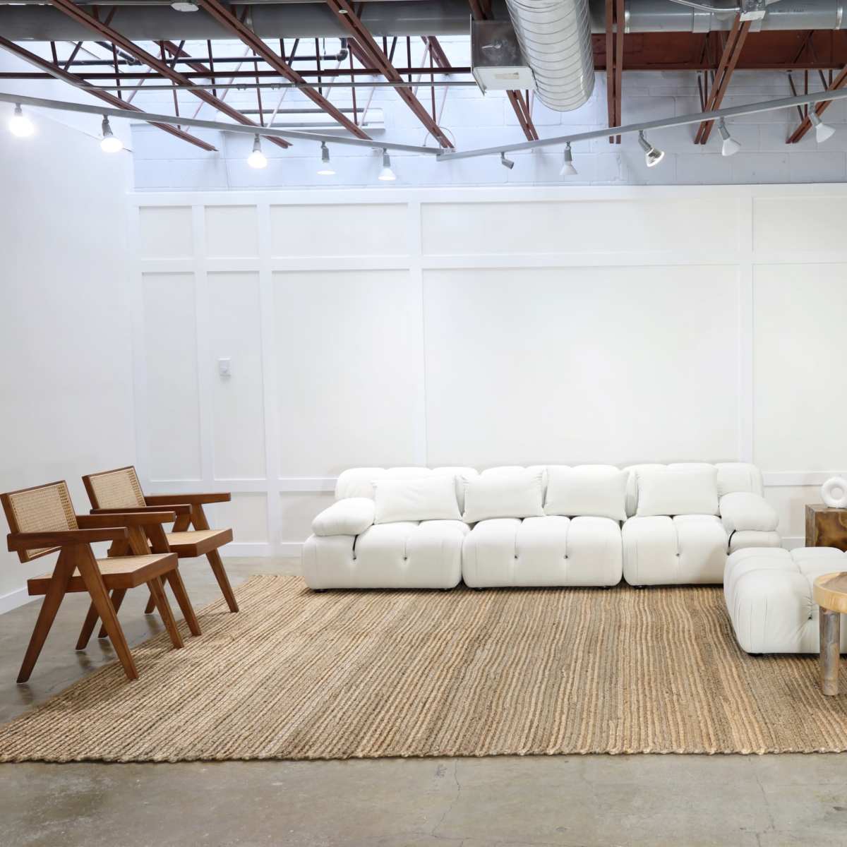 A modern living room with a white sectional sofa, a woven rug, a wooden side table, and a potted plant against a white wall.
