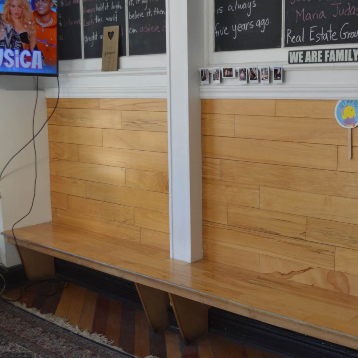 A wooden bench against a wall with a television mounted nearby and a bulletin board covered in photographs and notes.