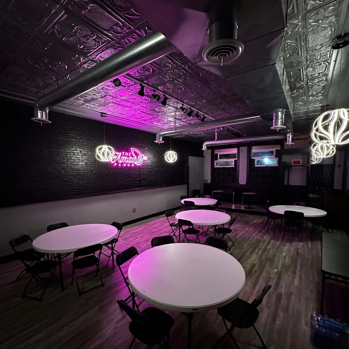 A dimly lit event space features several round tables surrounded by chairs, with pink neon lighting and decorative light fixtures on the ceiling.