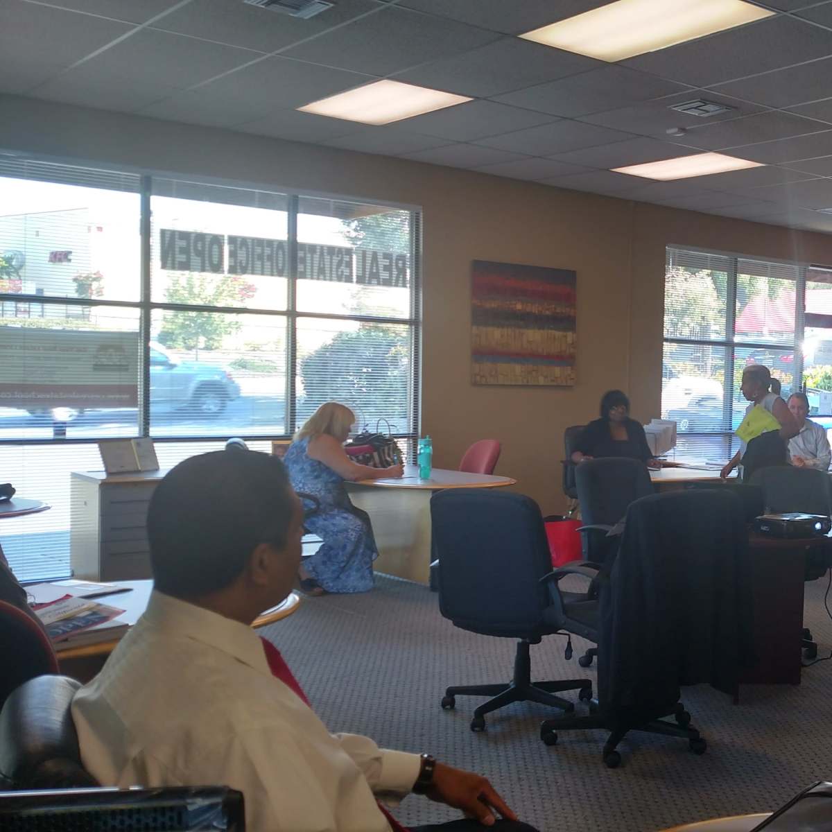 A group of people is gathered in a spacious office with desks and chairs, while one person stands speaking to the others.