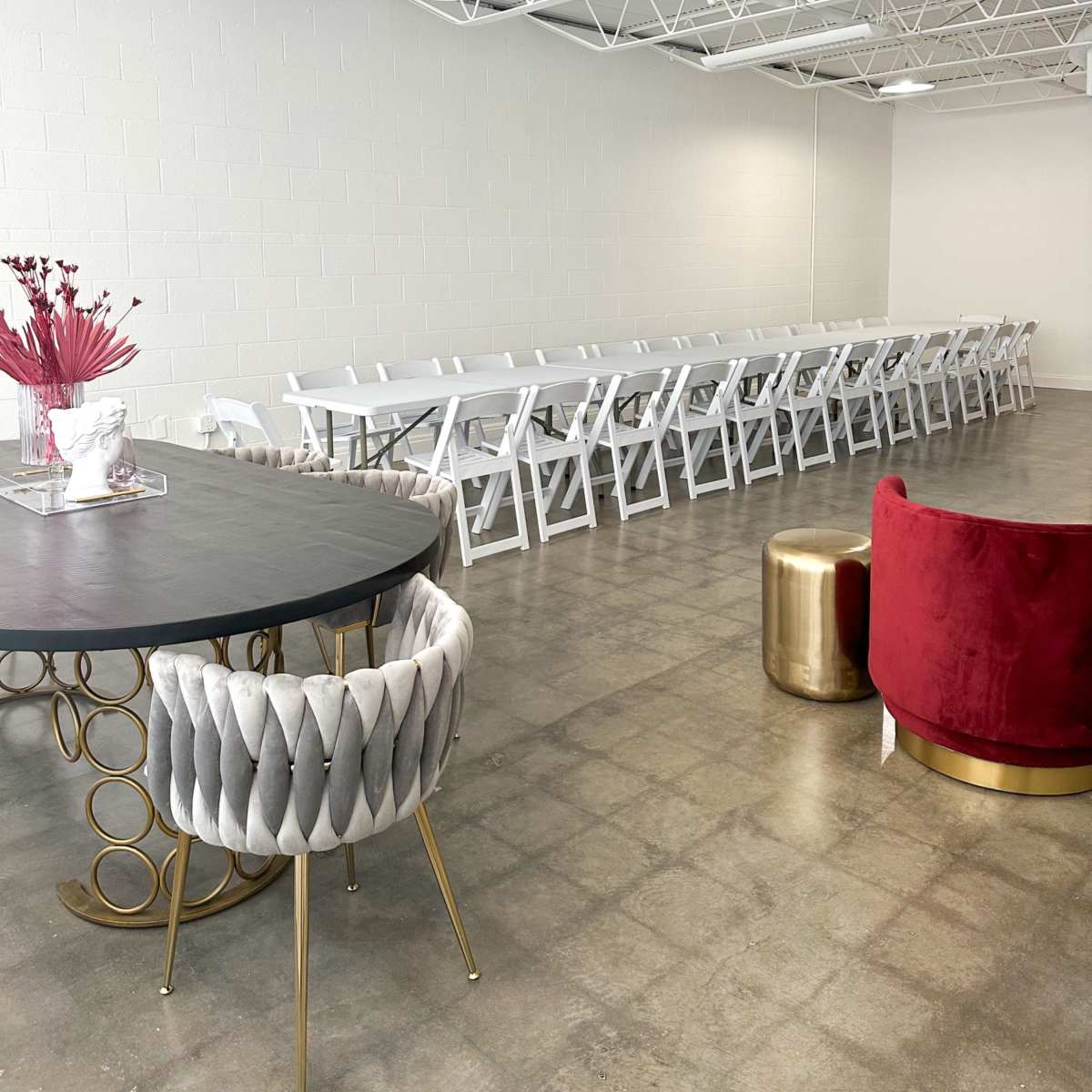 A spacious indoor venue featuring a long table lined with white folding chairs, a round table with a decorative centerpiece, and a red upholstered chair.