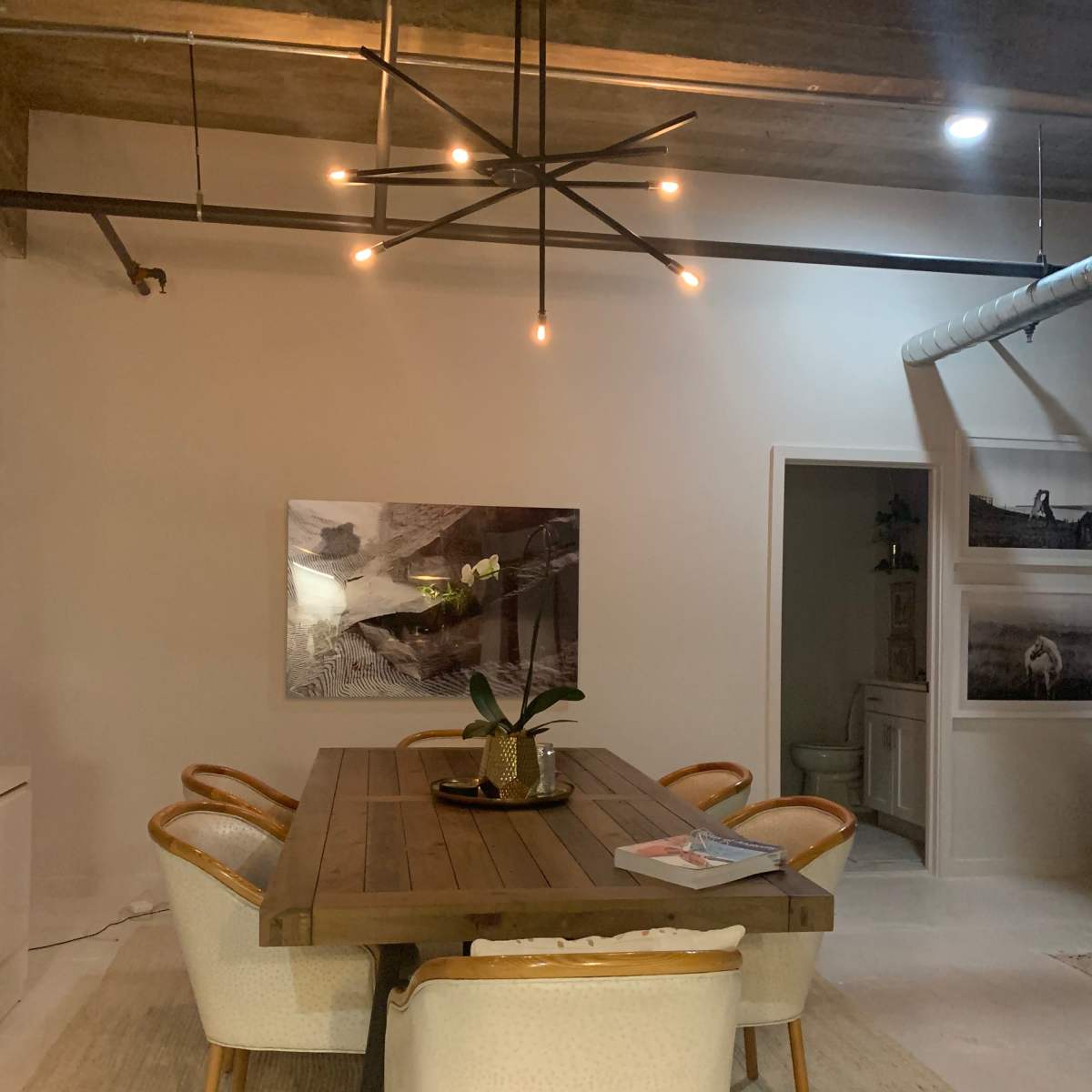 A dining area featuring a wooden table surrounded by upholstered chairs, illuminated by a modern chandelier, with artwork on the walls and a doorway leading to another room.