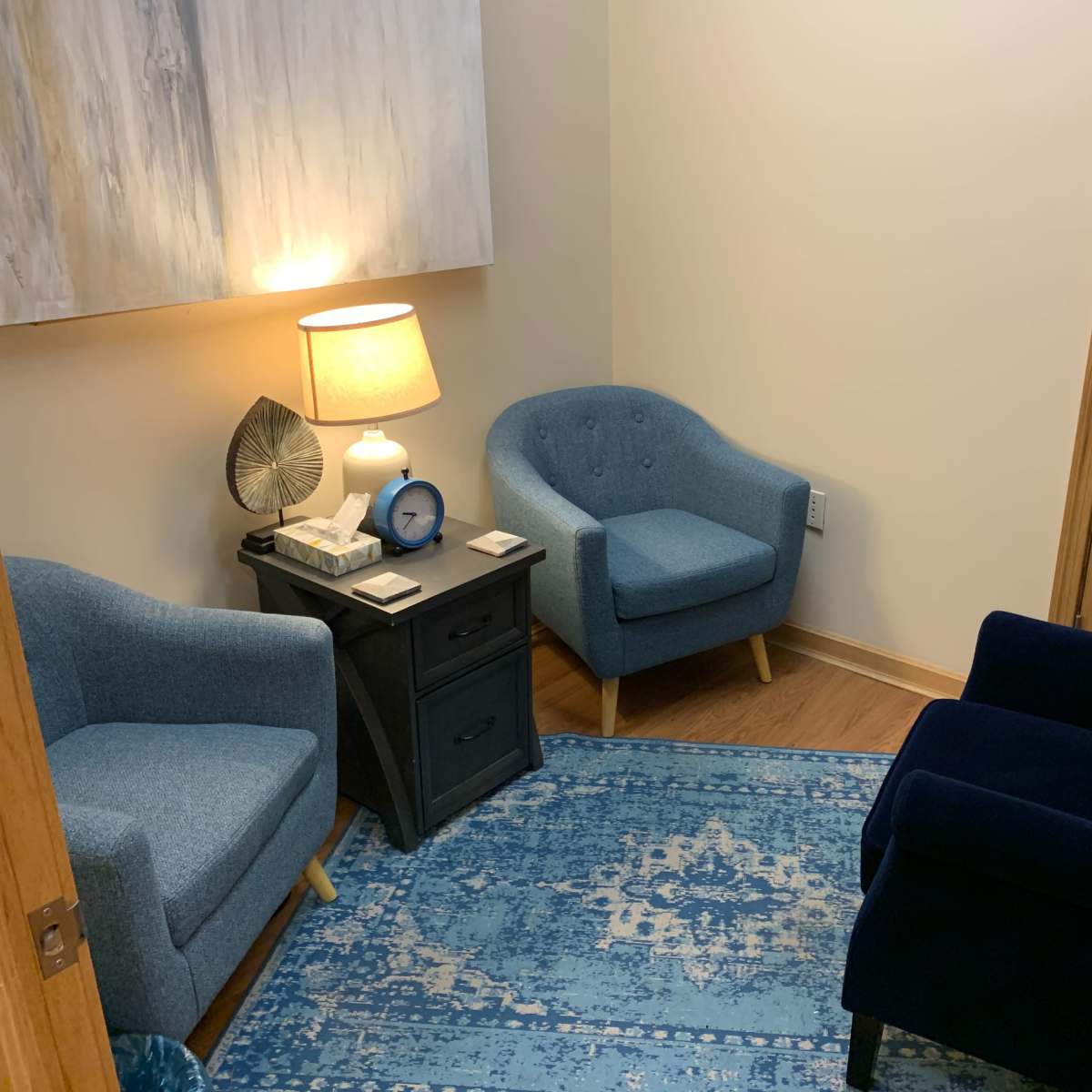 A small, softly lit room featuring two blue chairs, a dark nightstand with a lamp and clock, and a light blue area rug on a wooden floor.