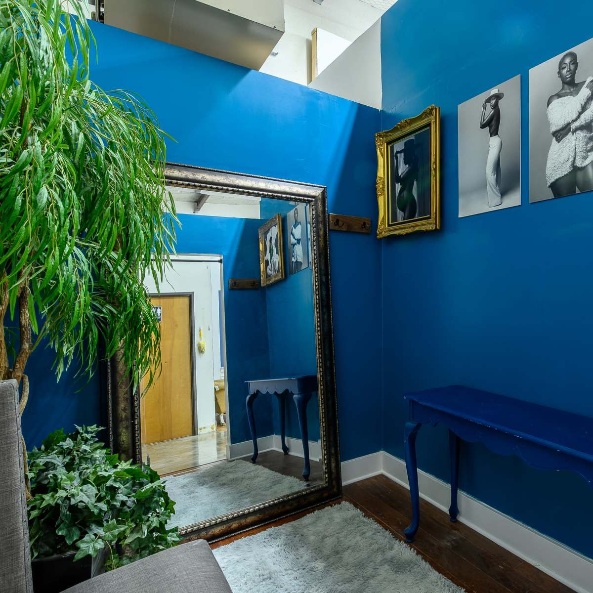 A small room with blue walls, featuring a large mirror, several framed black-and-white photographs on the wall, a blue console table, and green plants.