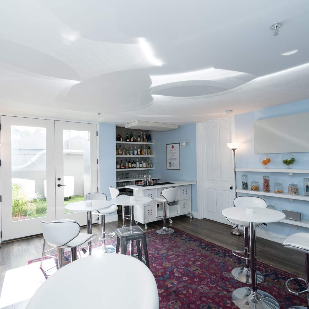 A modern lounge area with a bar and seating, featuring light blue walls, a patterned ceiling, and glass jars on shelves.