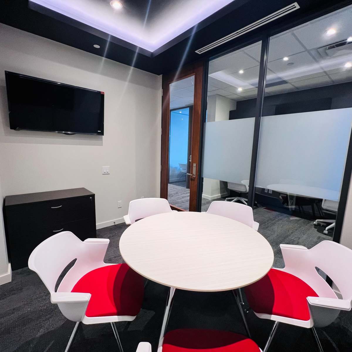 A small conference room features a round table with white and red chairs, a flat-screen TV on the wall, and glass partitions.