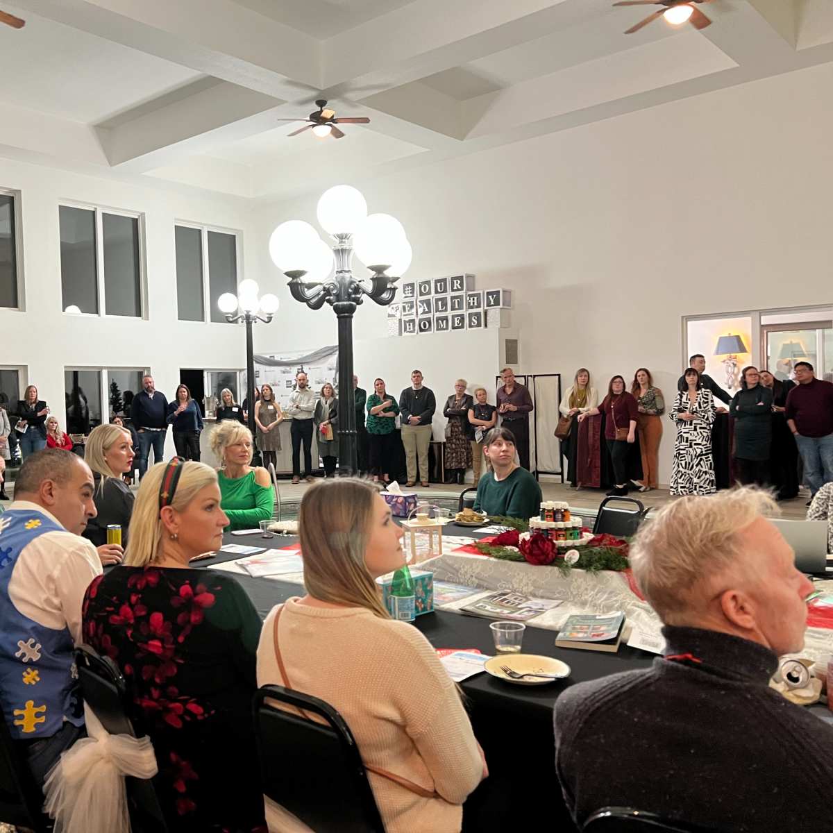 A large group of people is seated at a long table in a well-lit room, while others stand in the background listening to a speaker at a podium.