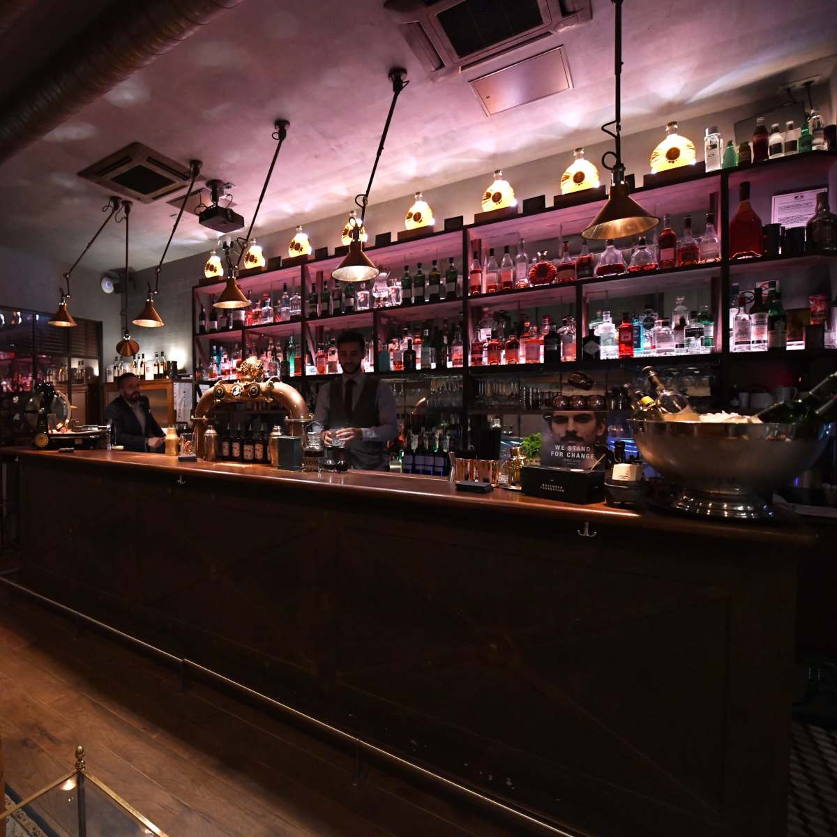 A dimly lit bar with a long wooden counter, shelves filled with various bottles, and leather chairs arranged around the space.