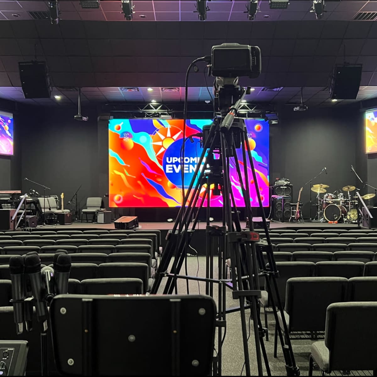 A staged interior of a venue set up for an event, featuring a large screen with colorful graphics, a sound and camera setup in the foreground, and empty rows of chairs facing the stage.