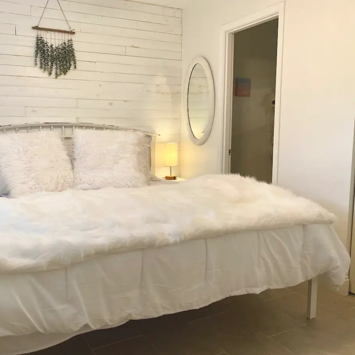 A white bed with fluffy pillows and a fur throw is positioned in a room with whitewashed walls and a round mirror.