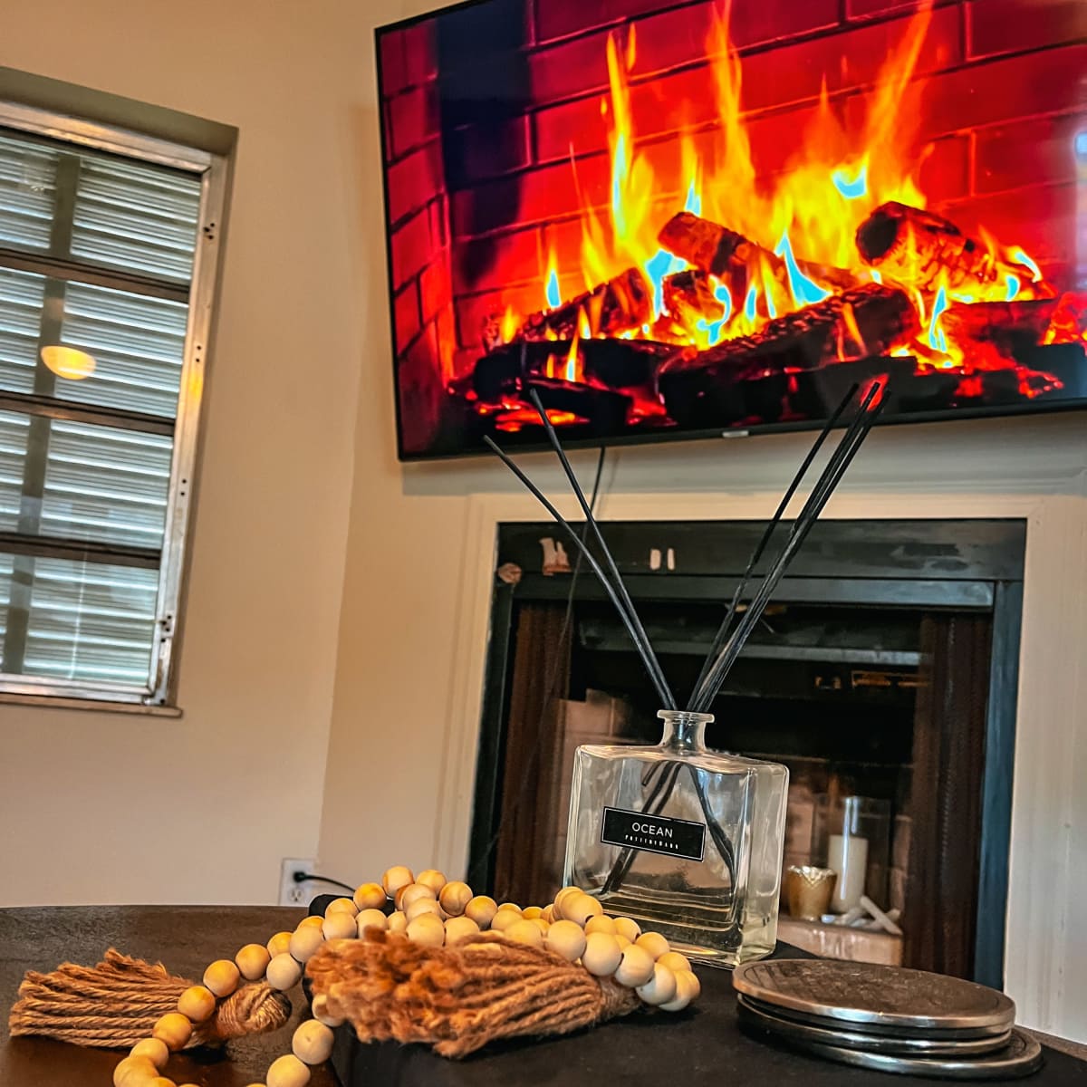 A decorative glass diffuser, wooden beads, and a closed book are placed on a table in front of a television displaying a fireplace scene.