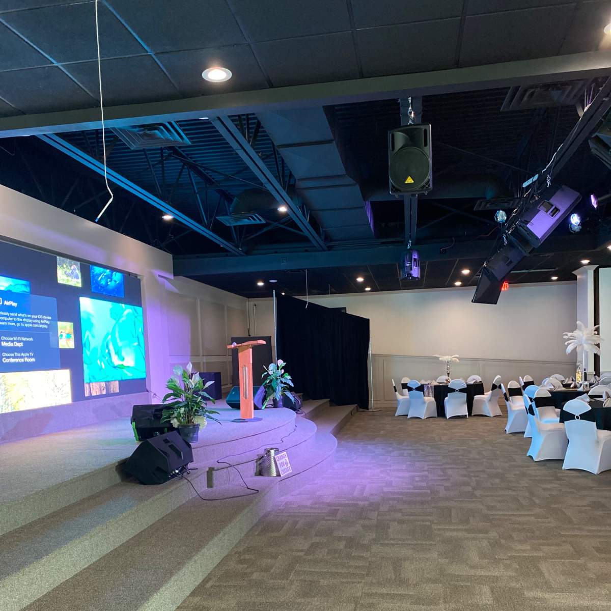 A stage with a backdrop featuring digital displays is set up in a banquet hall, with chairs arranged at tables nearby.