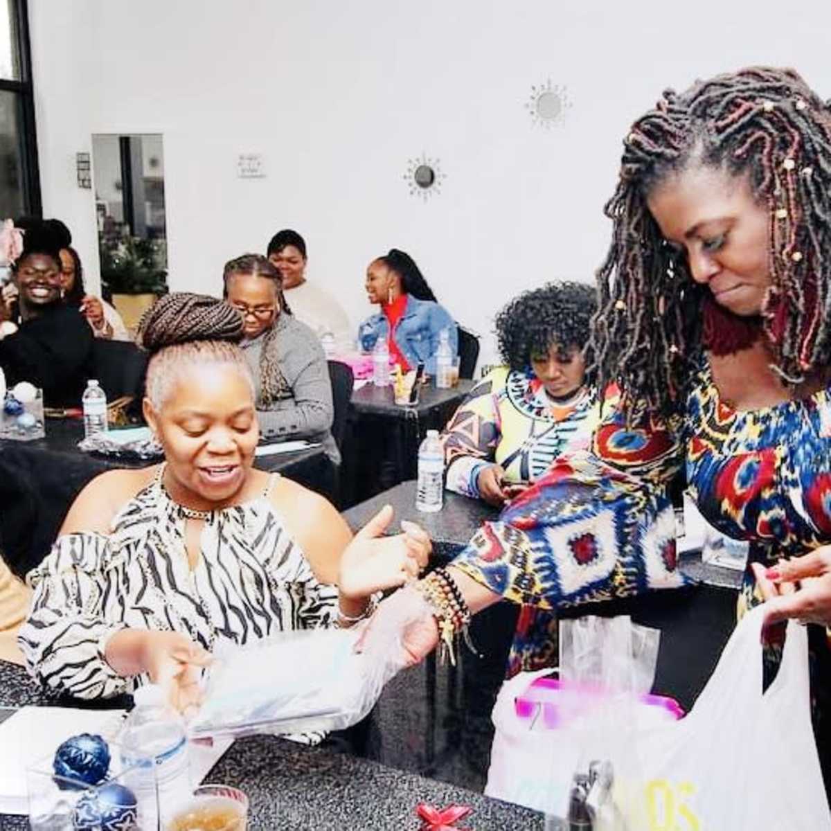 A group of people are gathered in a brightly lit room, engaging in conversation and exchanging items at a table covered with various supplies.
