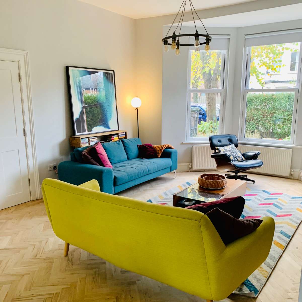 The living room features a blue sofa, a yellow armchair, a coffee table, and large windows allowing natural light to illuminate the space.