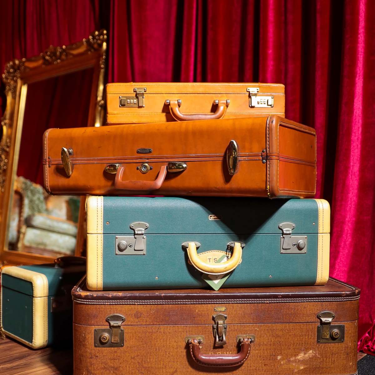 A stack of vintage suitcases in various colors is arranged next to a large ornate mirror against a red curtain backdrop.