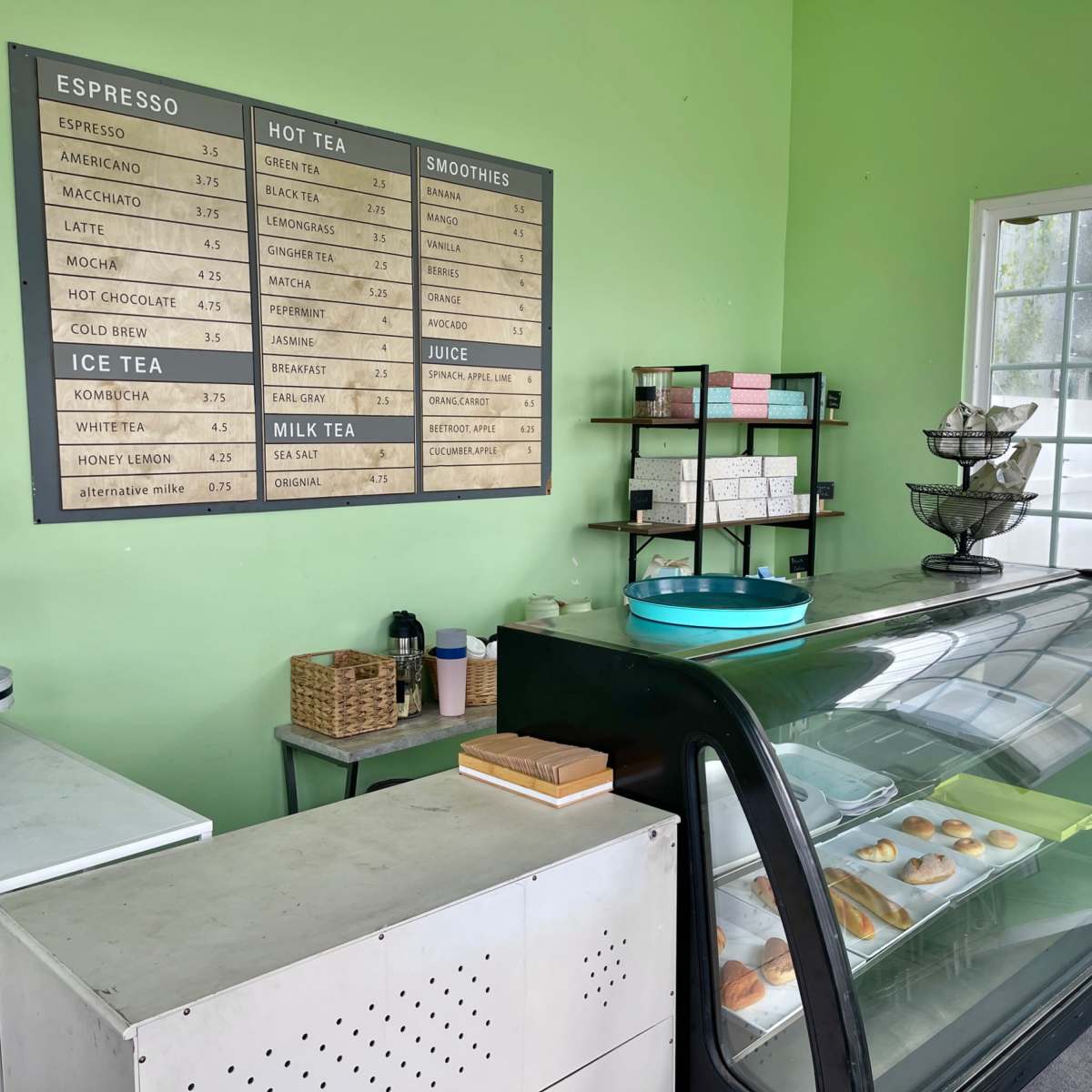 A small café counter with a display case holding pastries, a menu board on the wall, and green walls.
