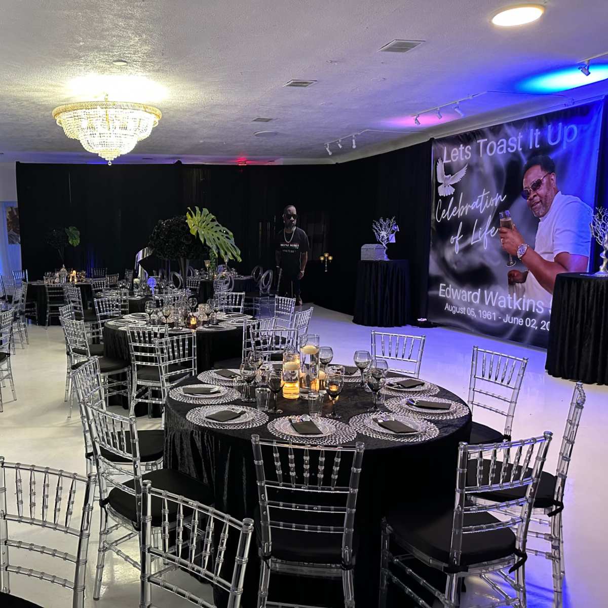 A banquet hall set up with round tables covered in black tablecloths, silver chiavari chairs, and decorative centerpieces, featuring a backdrop celebrating Edward Watkins with a large portrait.