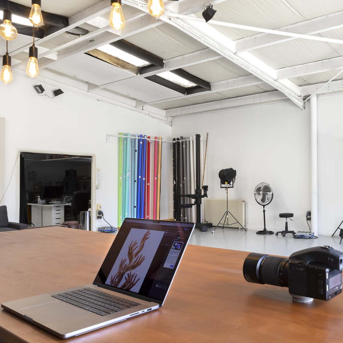 A spacious photography studio features a wooden table with a laptop and camera, surrounded by studio lighting equipment and backdrops.