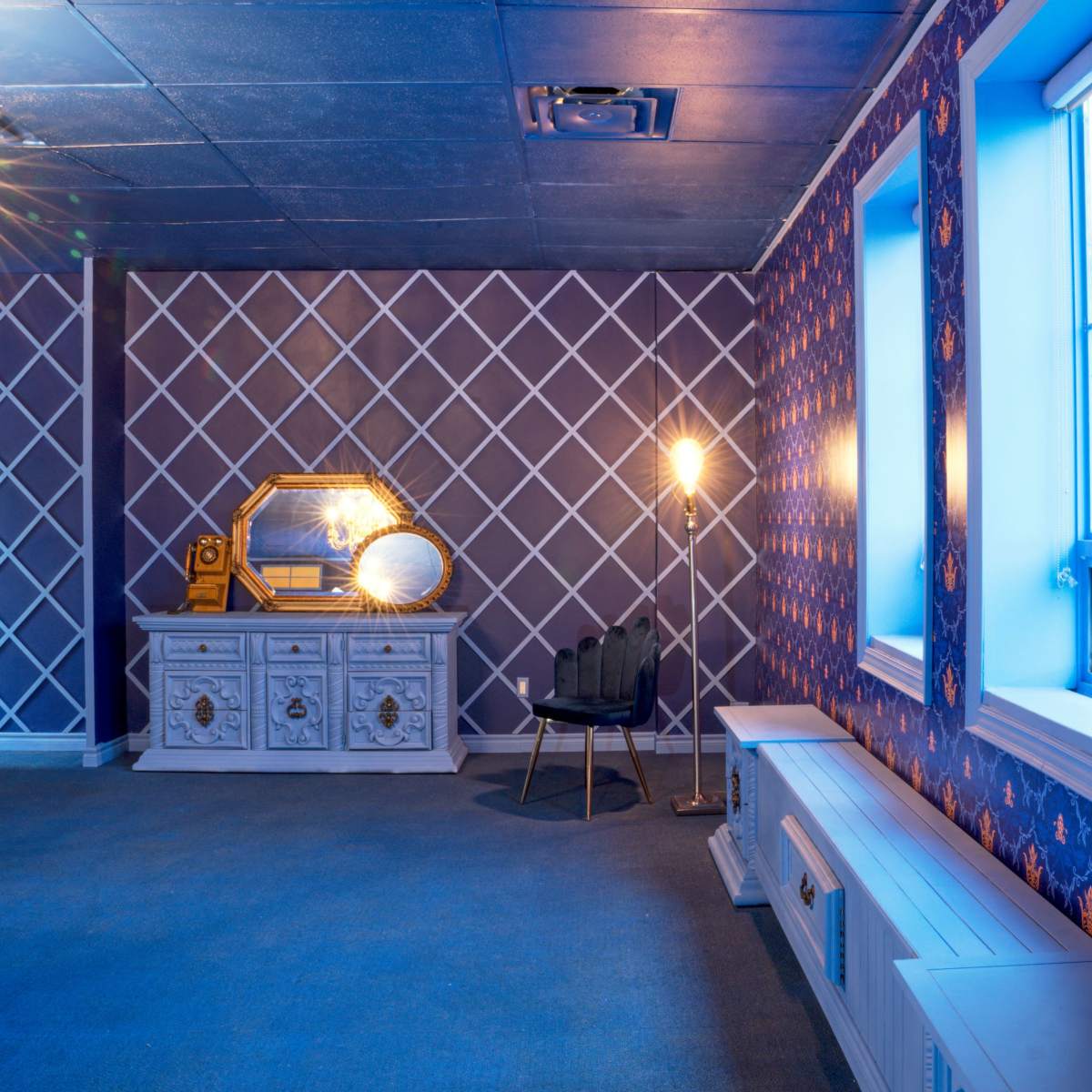 The room features a patterned wallpaper with a chandelier, a decorative mirror on a dresser, and a chair beside a lamp.