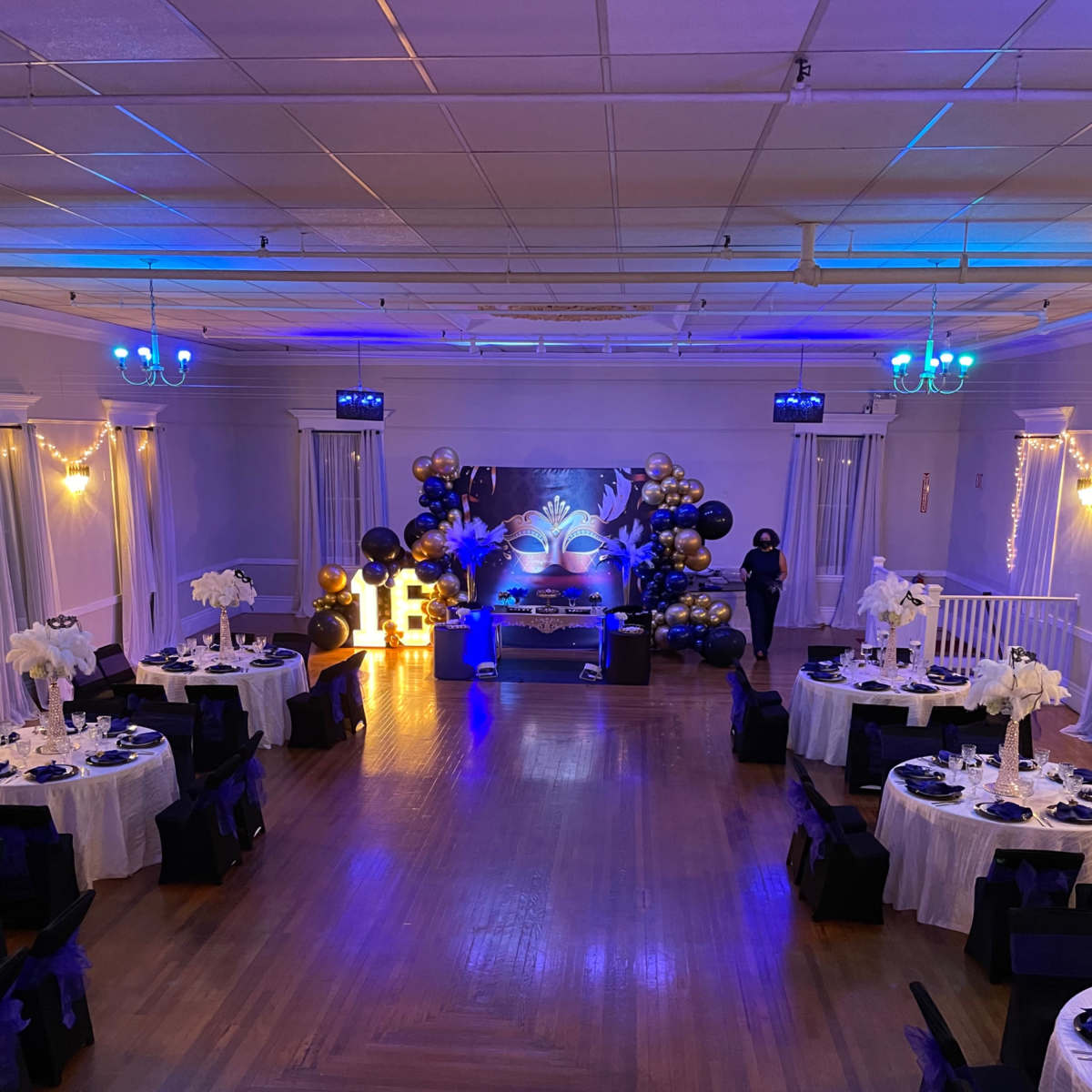 A decorated event hall with tables set for a celebration, featuring black and gold balloons, blue lighting, and floral centerpieces.