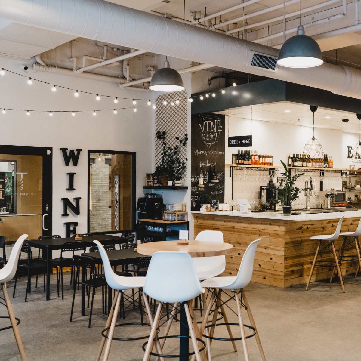 A modern, open café features a wooden bar, high tables with white stools, and shelves lined with wine bottles.