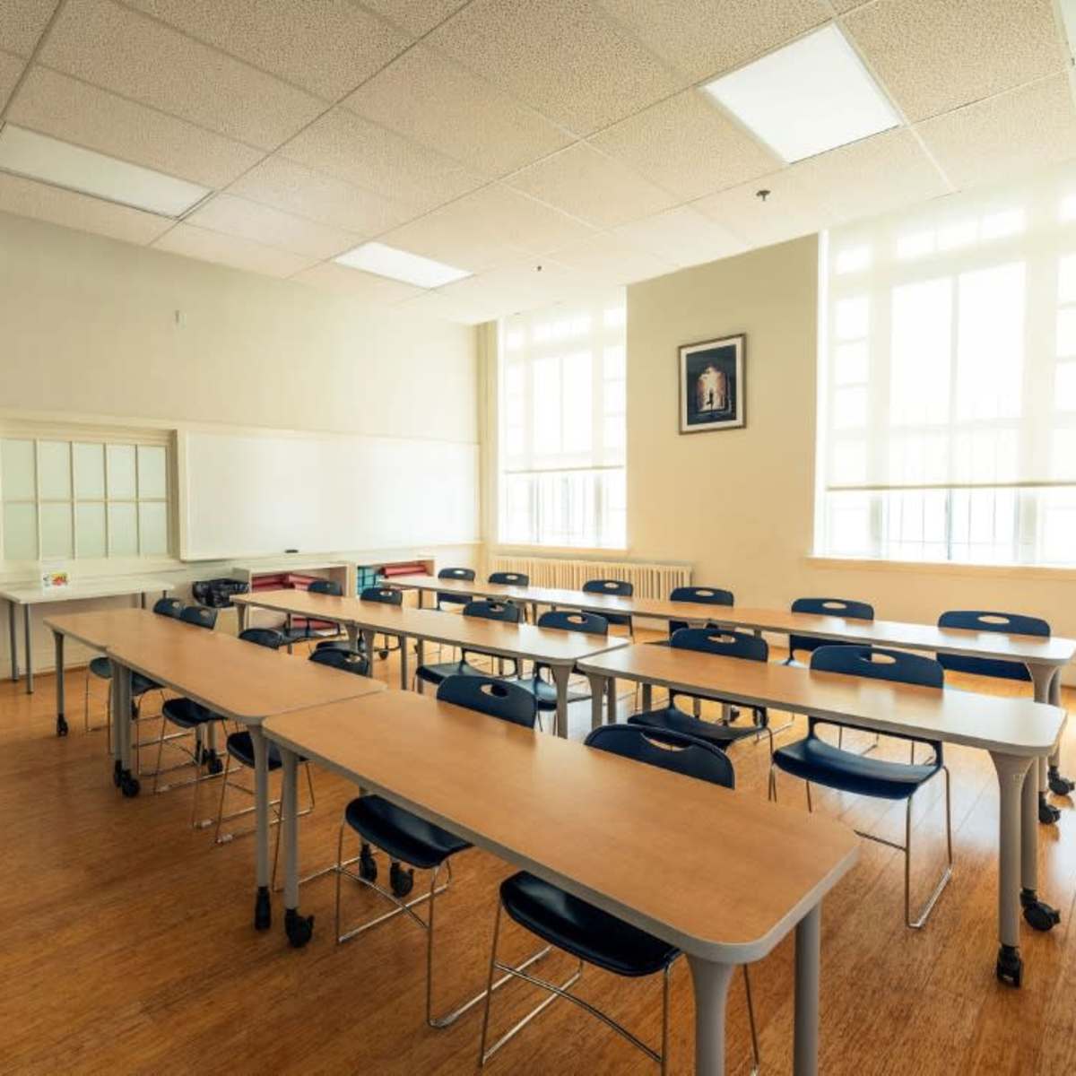 A classroom features several rows of tables and chairs, with large windows allowing natural light to enter.
