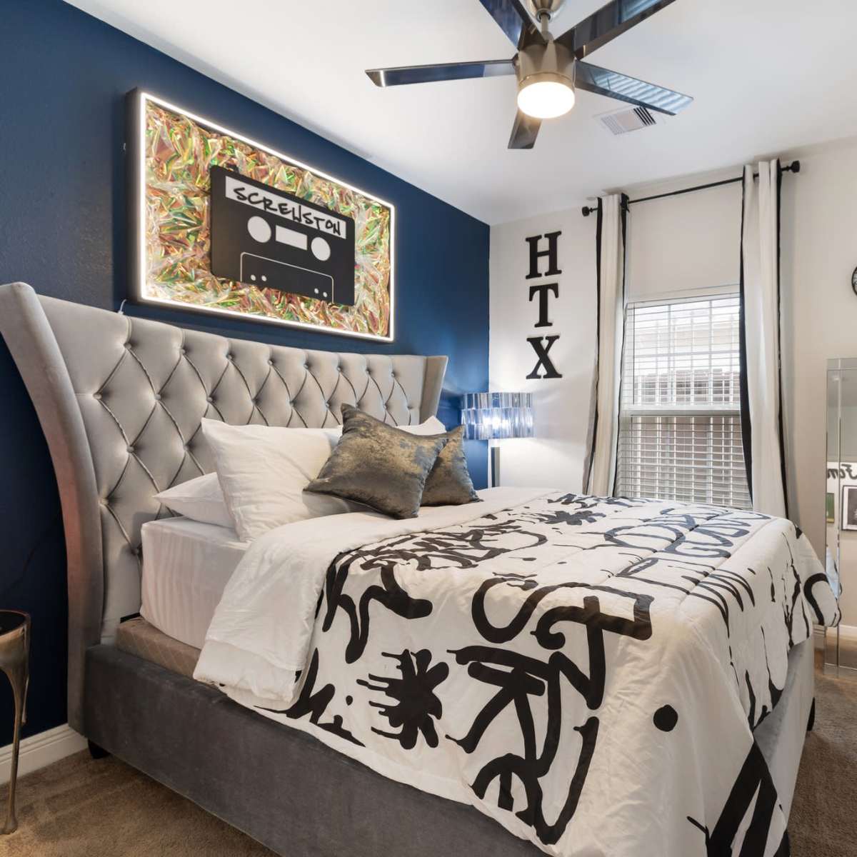 A neatly arranged bedroom featuring a tufted bed with a decorative black and white bedspread, a colorful artwork on the wall, and modern furnishings including a mirror and a wall-mounted TV.