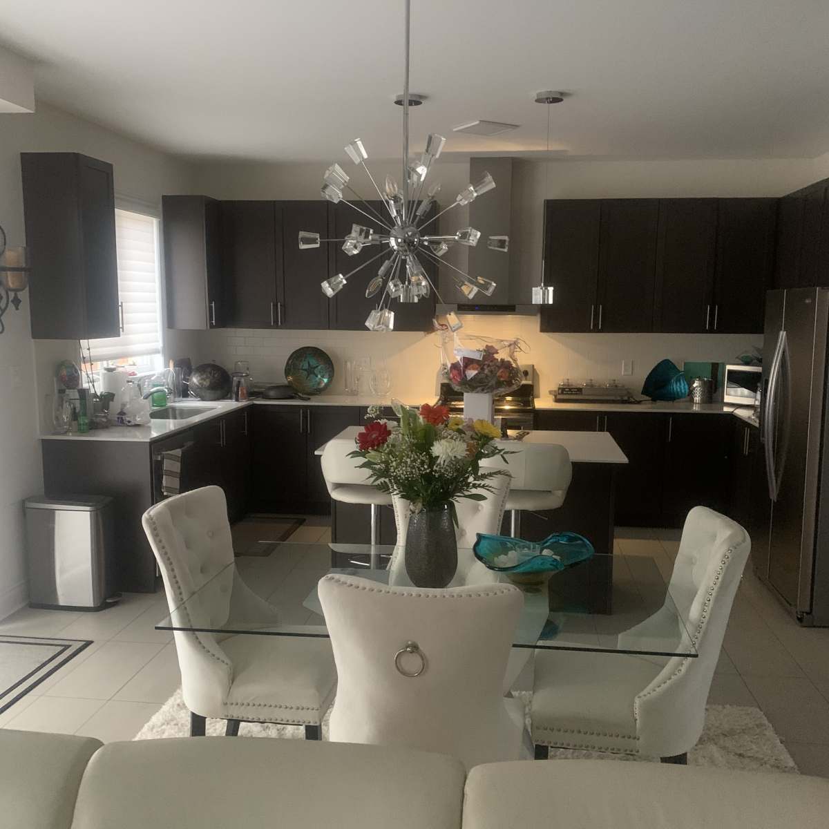 A modern kitchen features dark cabinets, a glass dining table surrounded by white chairs, and a contemporary chandelier.