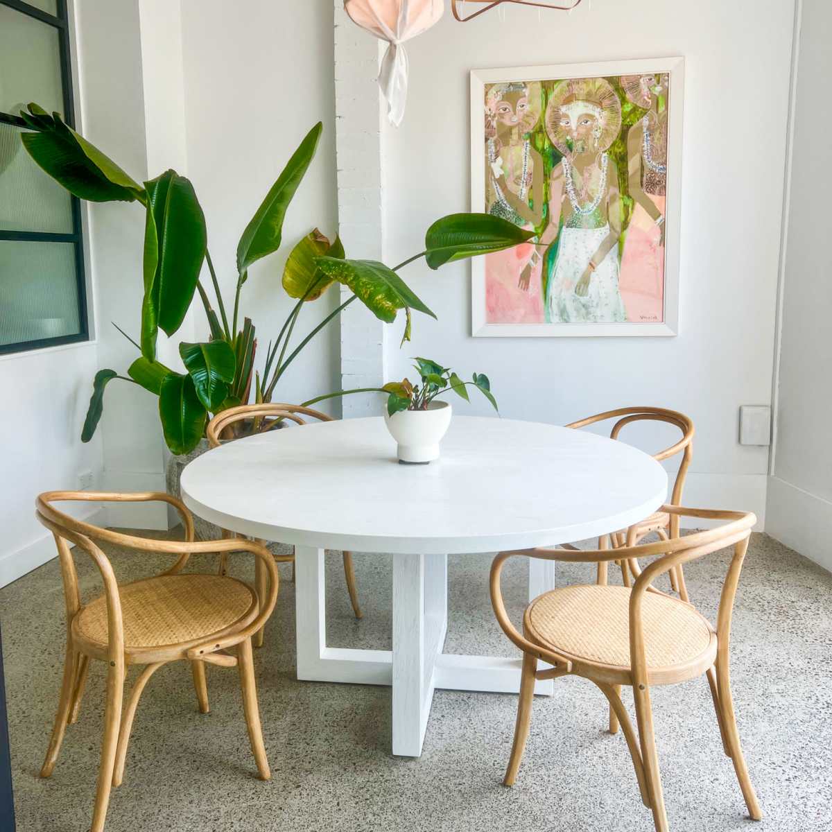 A round white dining table is surrounded by four rattan chairs, with a large plant and a colorful art piece on the wall in a well-lit room.