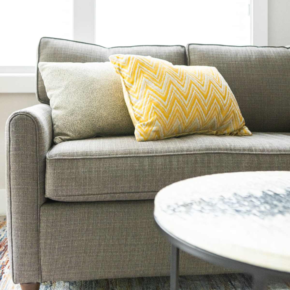 A gray sofa with patterned pillows is positioned next to a round table in a brightly lit room.