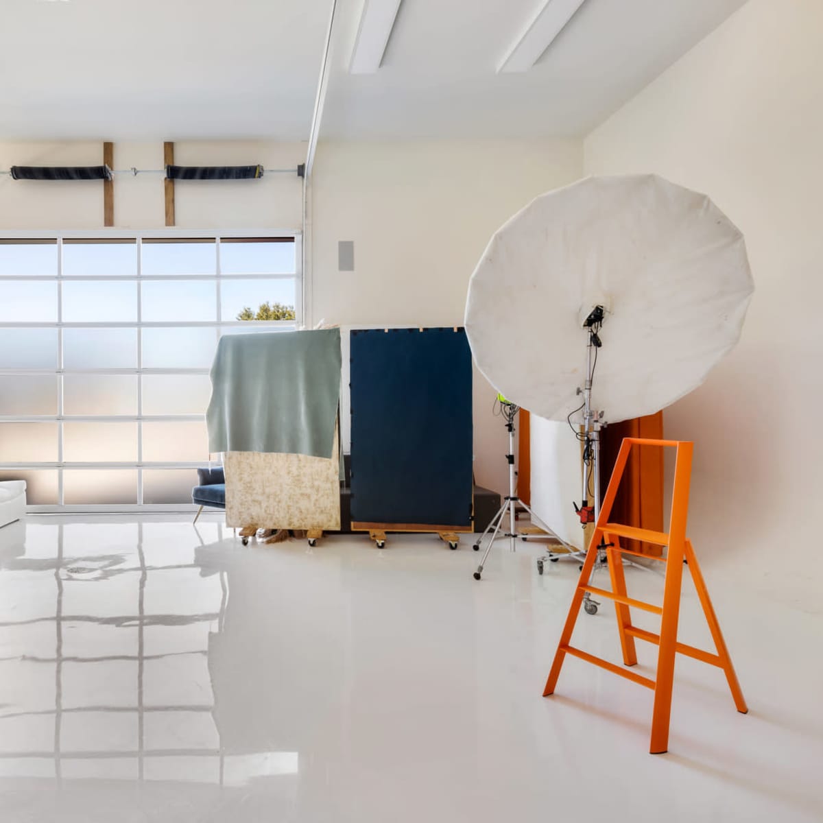 A bright studio space featuring a white couch, a wooden coffee table, a large reflective window, a fabric backdrop, a photography umbrella, and an orange director's chair.