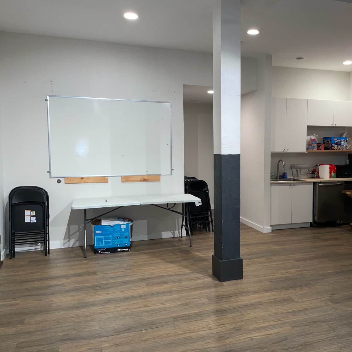 A spacious interior room with a kitchenette, a whiteboard on the wall, and several tables and chairs arranged for use.