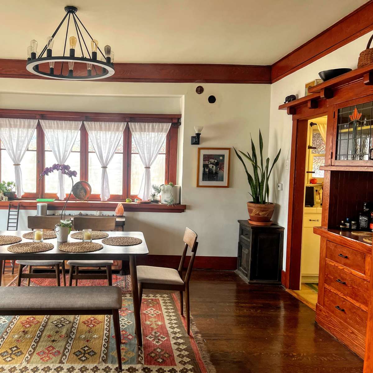 A dining area with a wooden table surrounded by chairs, large windows adorned with white curtains, and decorative plants along the window ledge.