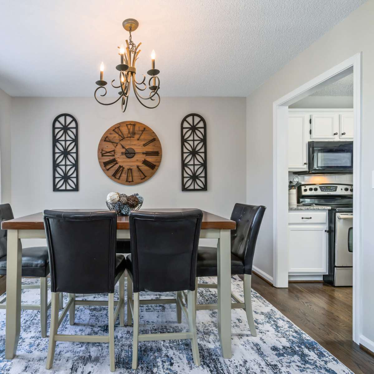 A dining area features a wooden table surrounded by black chairs, with a large clock on the wall and a view of the kitchen in the background.
