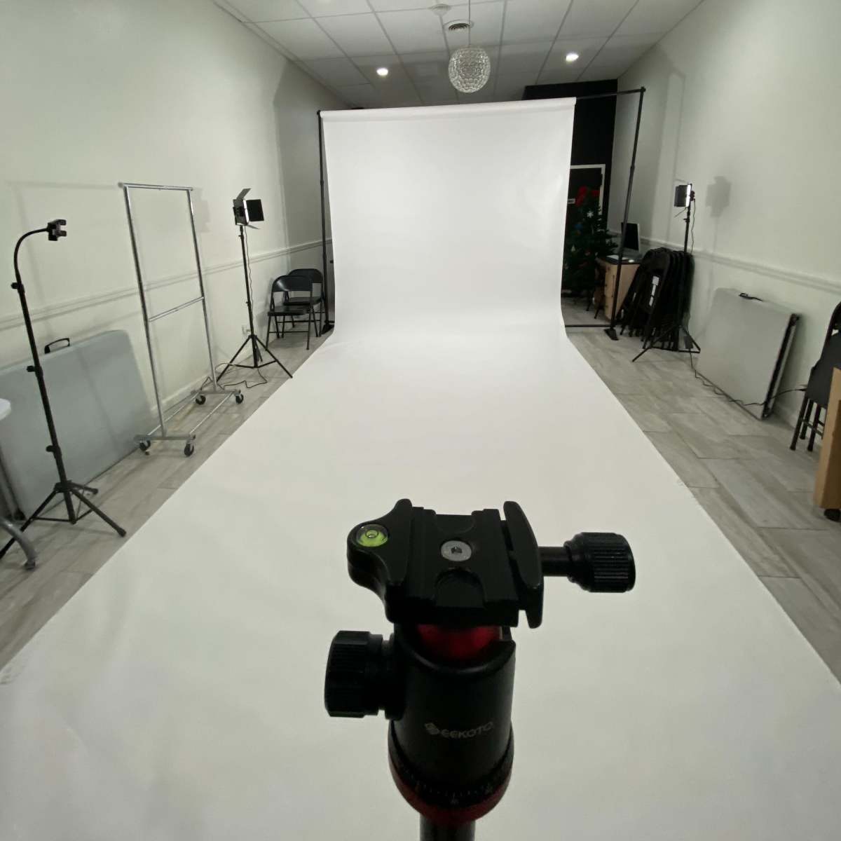A photography studio is set up with a white backdrop, tripod in the foreground, and various lighting equipment arranged around the room.