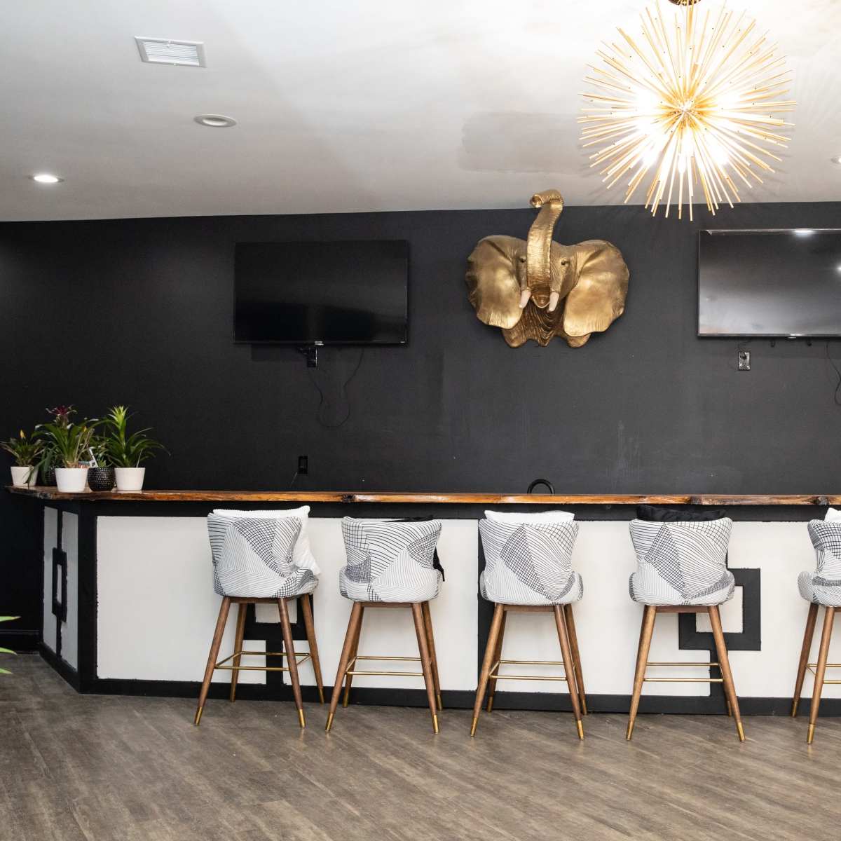 A modern bar area with a wooden counter, five stools, two wall-mounted televisions, and a decorative wall piece resembling an elephant's head.