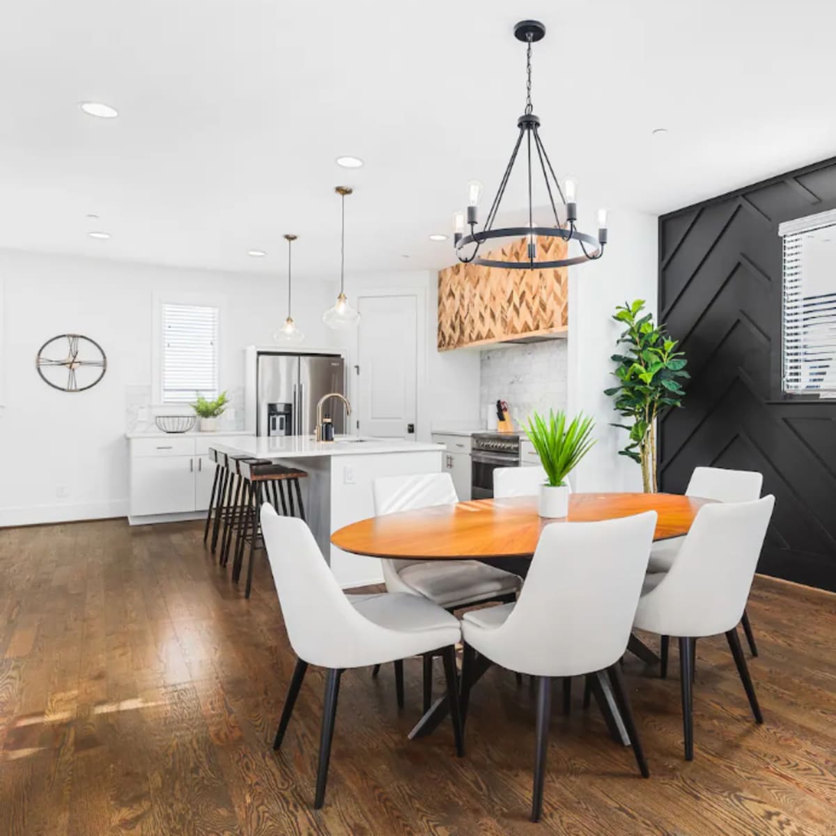 A modern dining area features a round wooden table surrounded by white chairs, with a sleek kitchen in the background and a black accent wall.