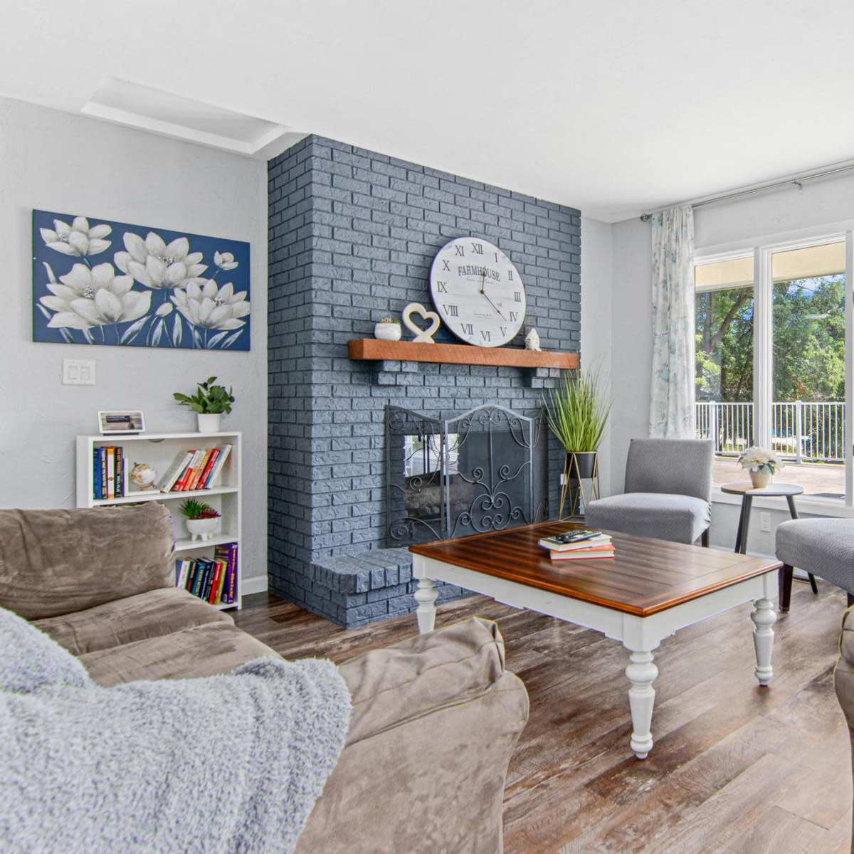 A cozy living room featuring a blue brick fireplace, a wooden coffee table, and two gray chairs, with large windows overlooking an outdoor area.