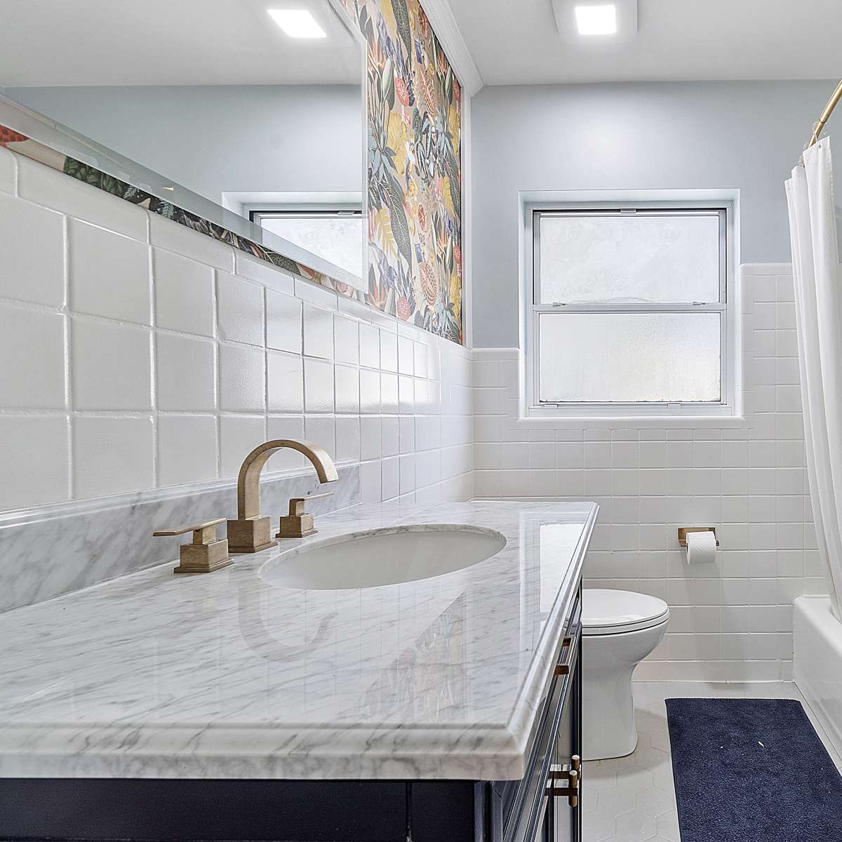 A bathroom featuring a marble countertop, a white tiled bathtub with a shower curtain, and a window allowing natural light.