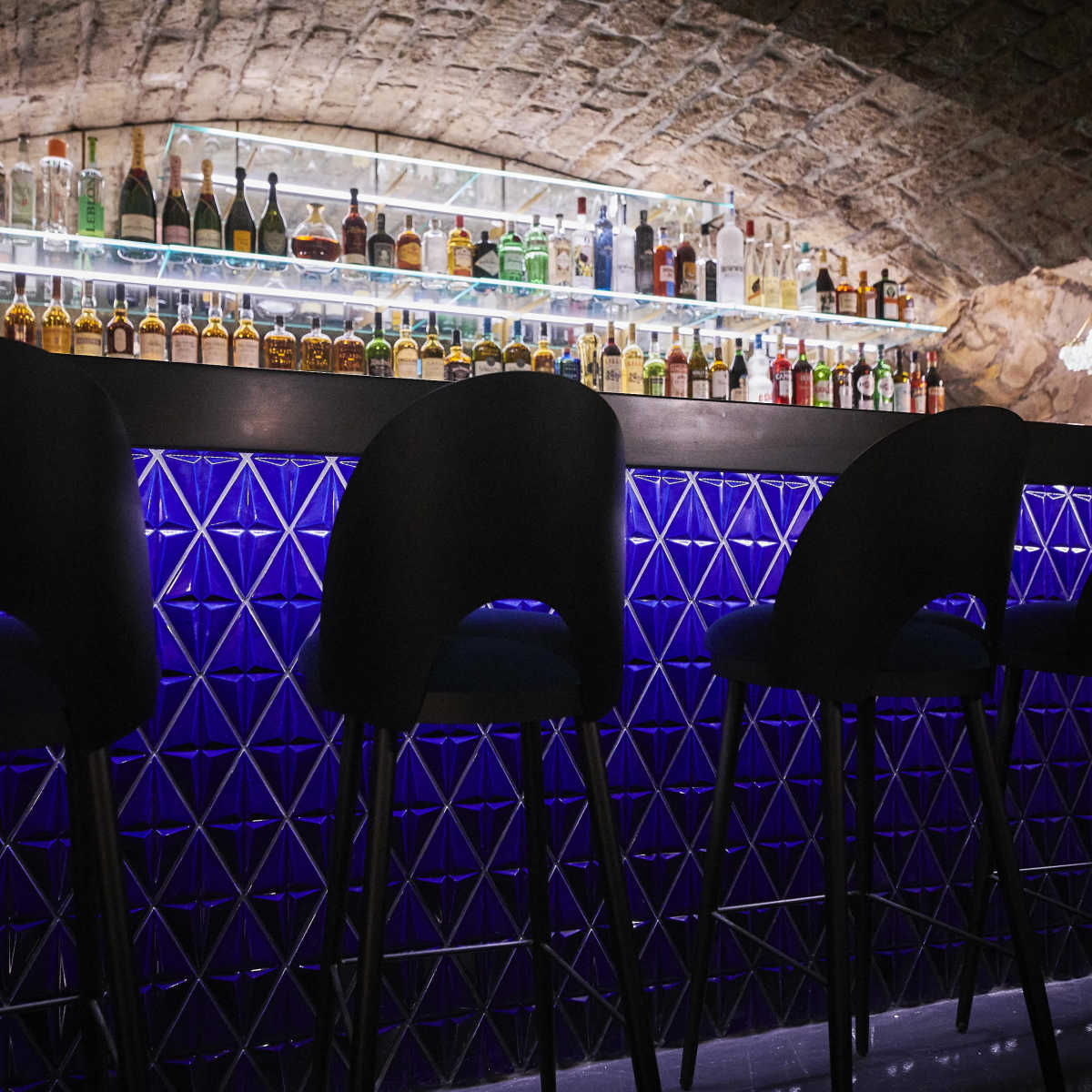 A dimly lit bar features a backlit shelf stocked with various bottles of liquor, accompanied by high black stools arranged in front of a blue geometric-patterned bar surface.