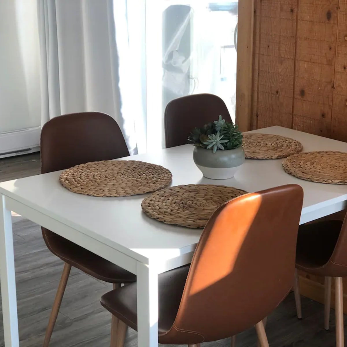 A white dining table is set with four brown chairs and round woven placemats, accompanied by a small potted plant at the center.