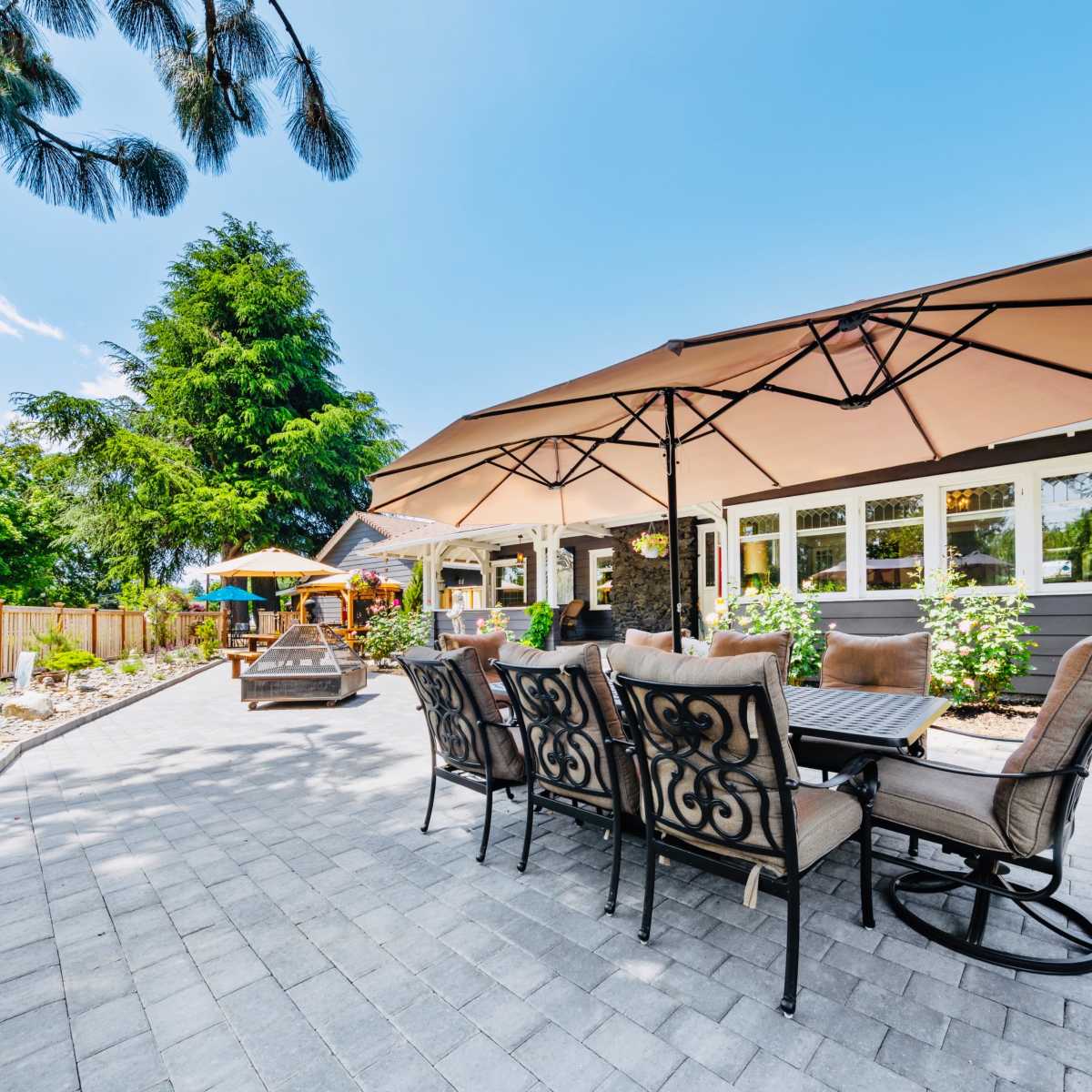 A patio area features a large dining set under beige umbrellas, surrounded by greenery and landscaped stones.