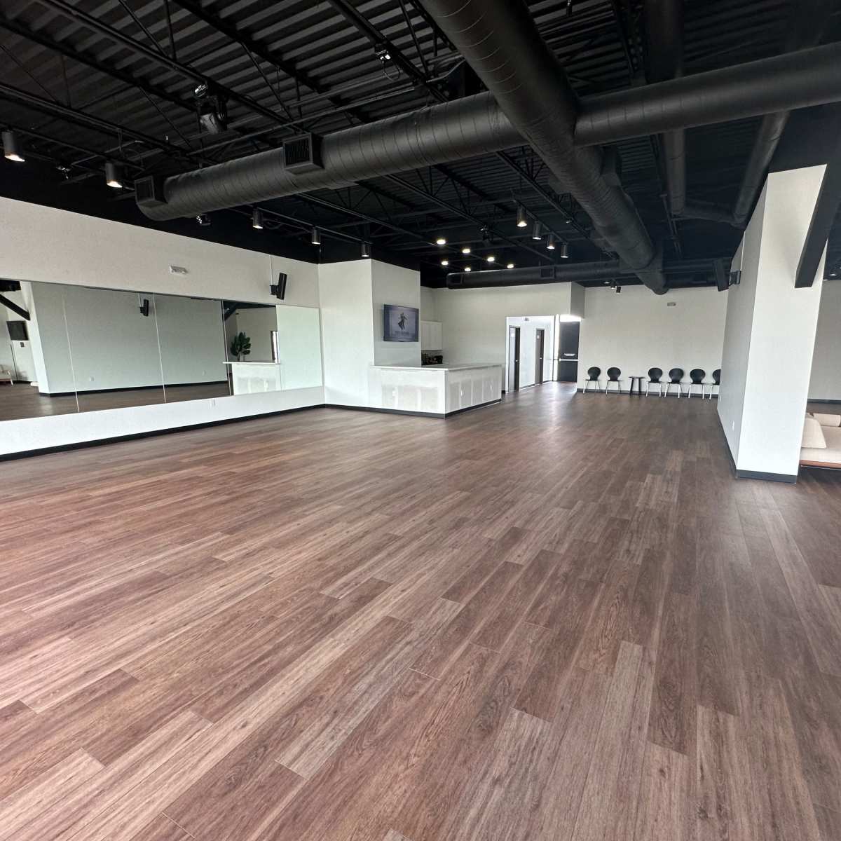 A spacious, well-lit studio with wooden flooring, large mirrors on one wall, and a seating area in the corner.